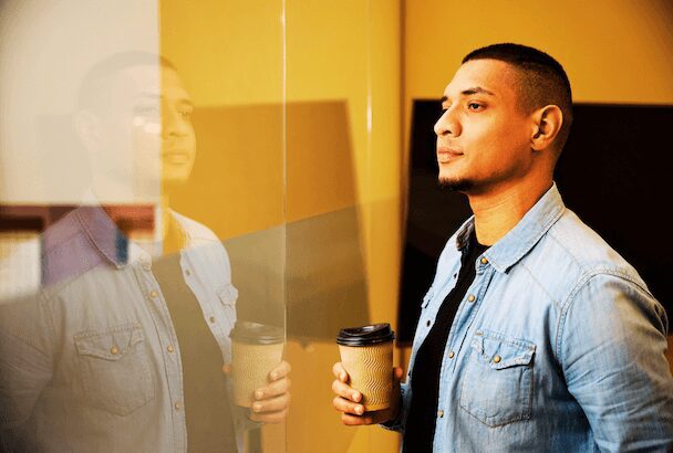 A young man holding coffee, wearing denim jacket, standing in warm light contemplating his core values..