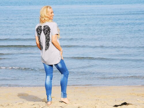 Dr. Madisen Harper is on the beach wearing a winged t-shirt, embodying freedom and tranquility by the sea.