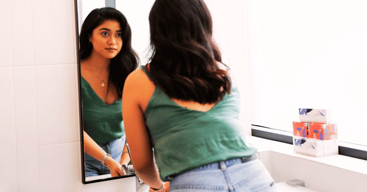 Young woman in green tank top contemplating her reflection in modern, bright bathroom.