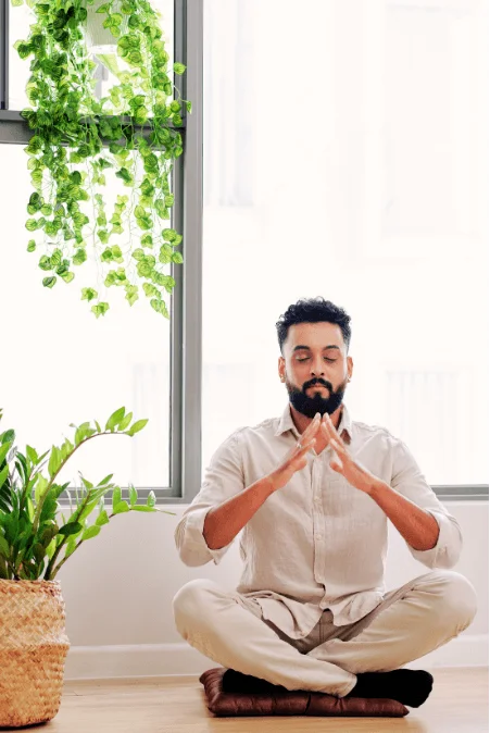 Man meditating on floor with greenery, sunlight, and serene atmosphere.